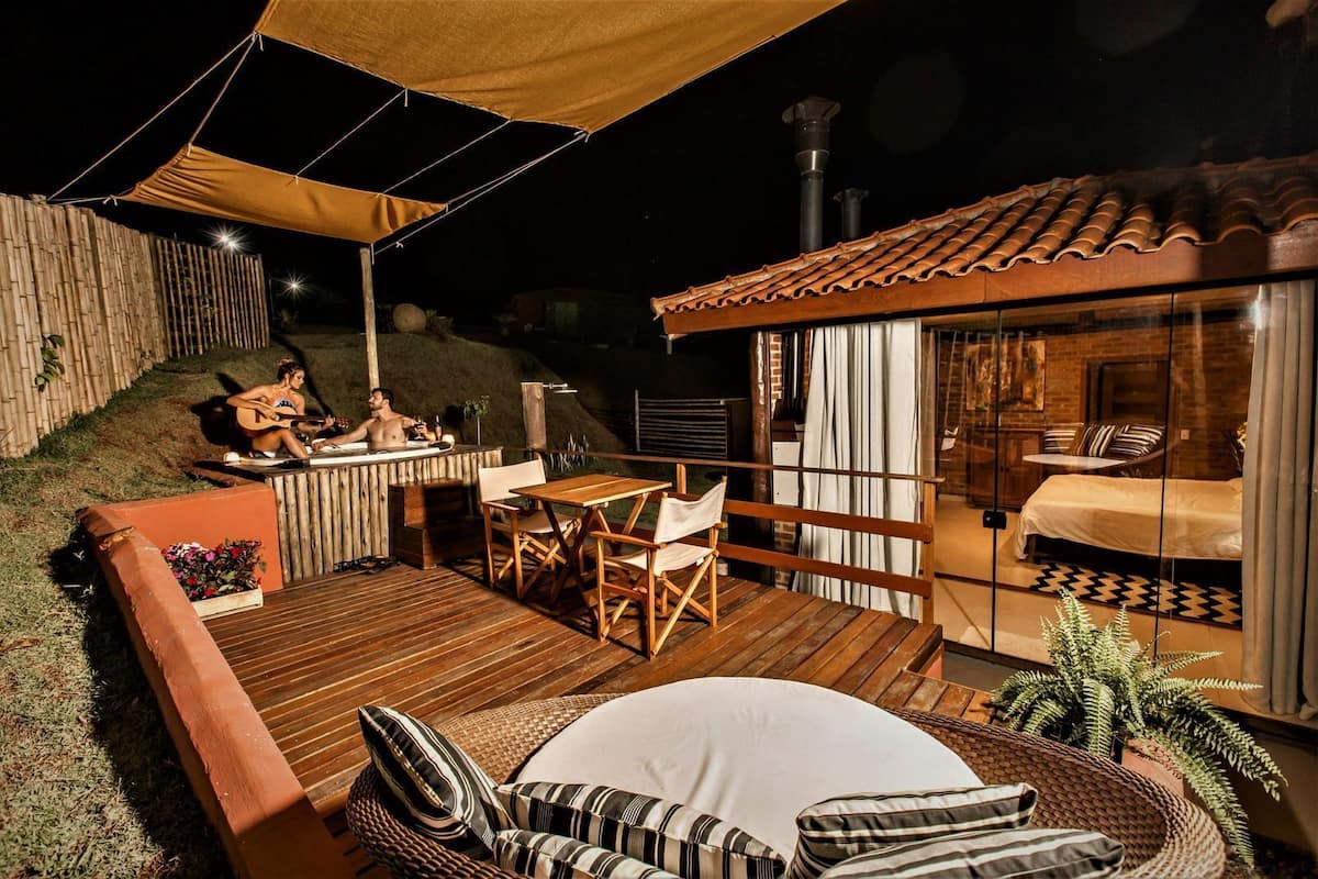 A cozy outdoor area with a wooden deck, a seating area with cushions, and a glass-enclosed room with a bed and a table.
