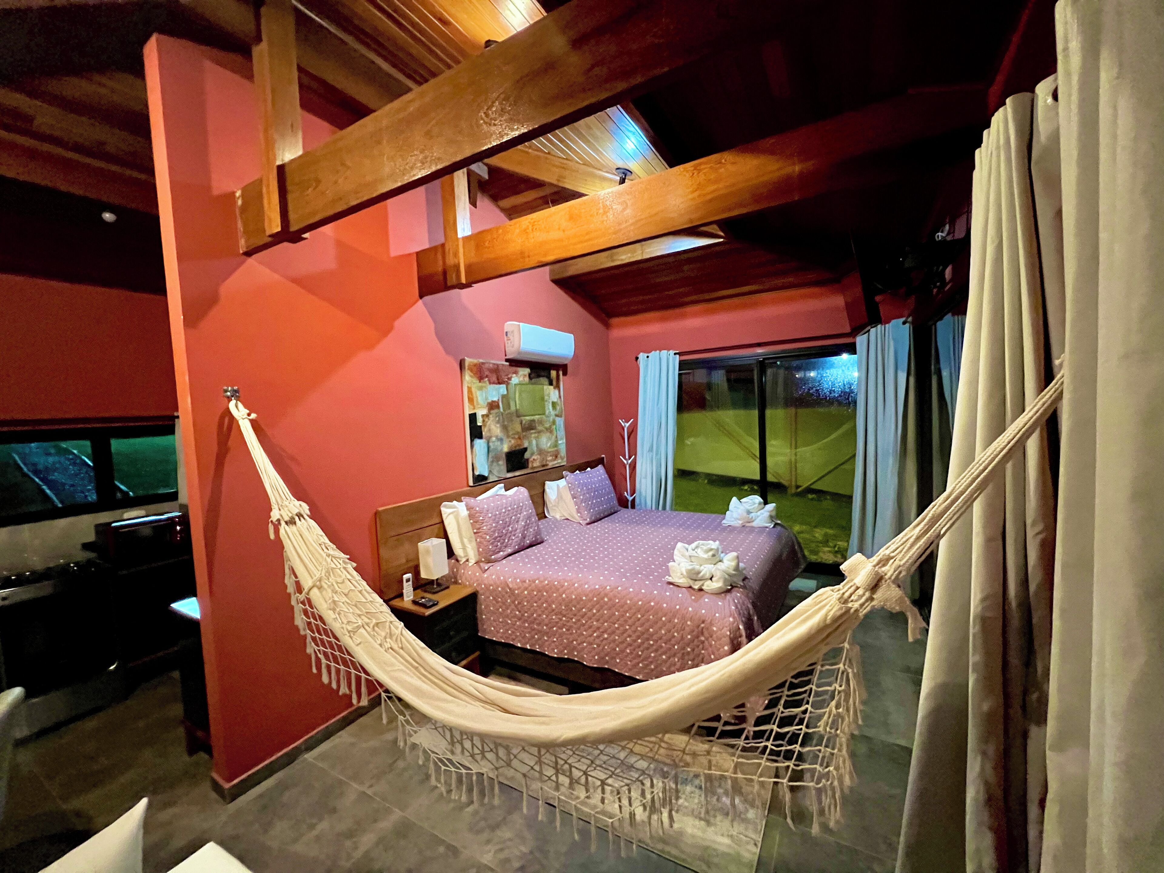 Um quarto com uma cama, uma rede, uma mesinha de cabeceira, um espelho e uma janela com cortinas.
