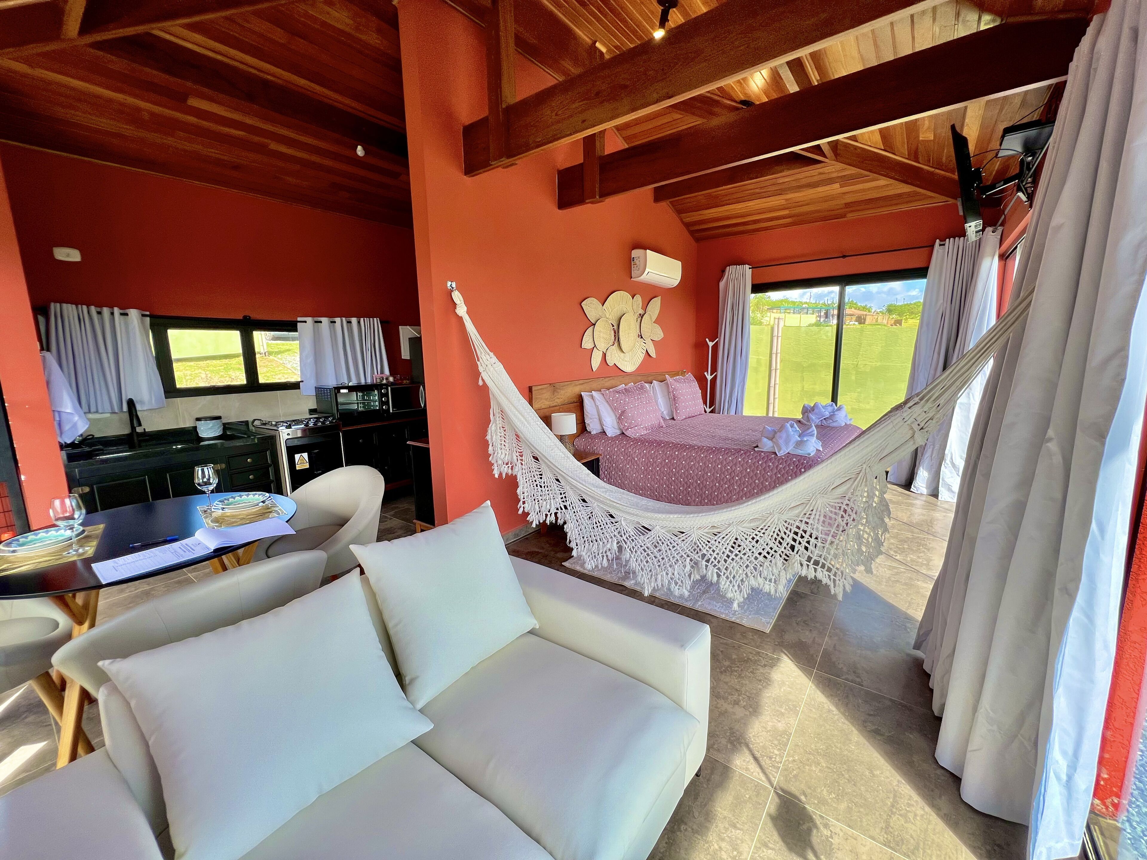 A room with a hammock bed, a dining area with a table and chairs, a kitchen area, and large windows with curtains.