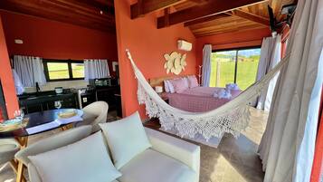 A room with a hammock bed, a dining area with a table and chairs, a kitchen area, and large windows with curtains.