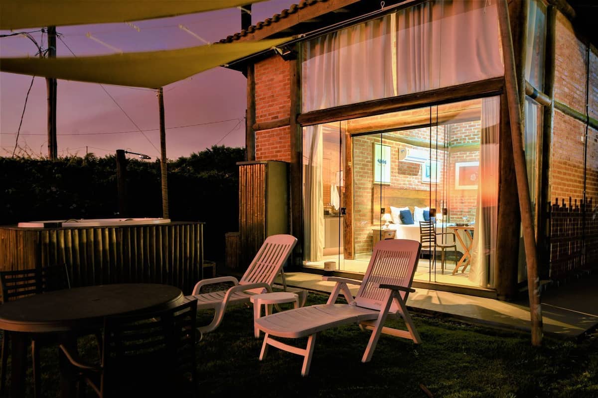 An outdoor patio area with a table and two chairs, a brick building with a glass door leading to a room, and a wooden fence in the background.