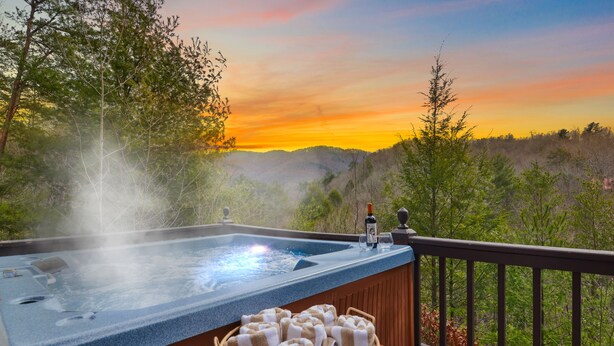 Ein Whirlpool mit einer Holzdielen-Terrasse und einem Korbwäscheschrank voller Handtücher, mit Blick auf eine bewaldete Landschaft bei Sonnenuntergang.