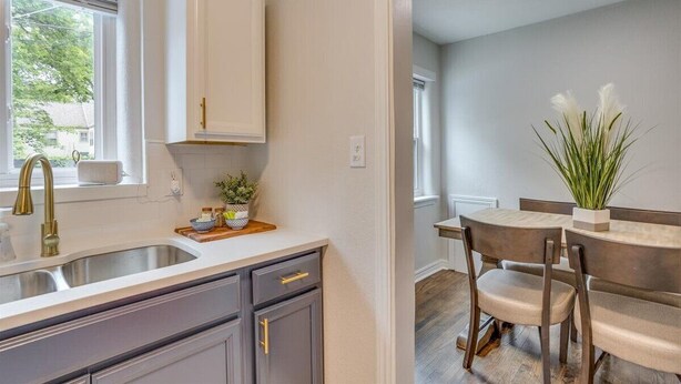 A modern kitchen with a white countertop, a gold faucet, and a window with blinds. There is a dining area with a wooden table and chairs, and a potted plant on the table. - Nearby Wesley. Pickle Ball + Family Fun! (Wichita)