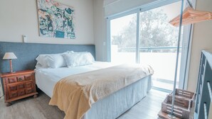 Un dormitorio con cama, mesita de noche, lámpara, ventana y vistas al exterior.