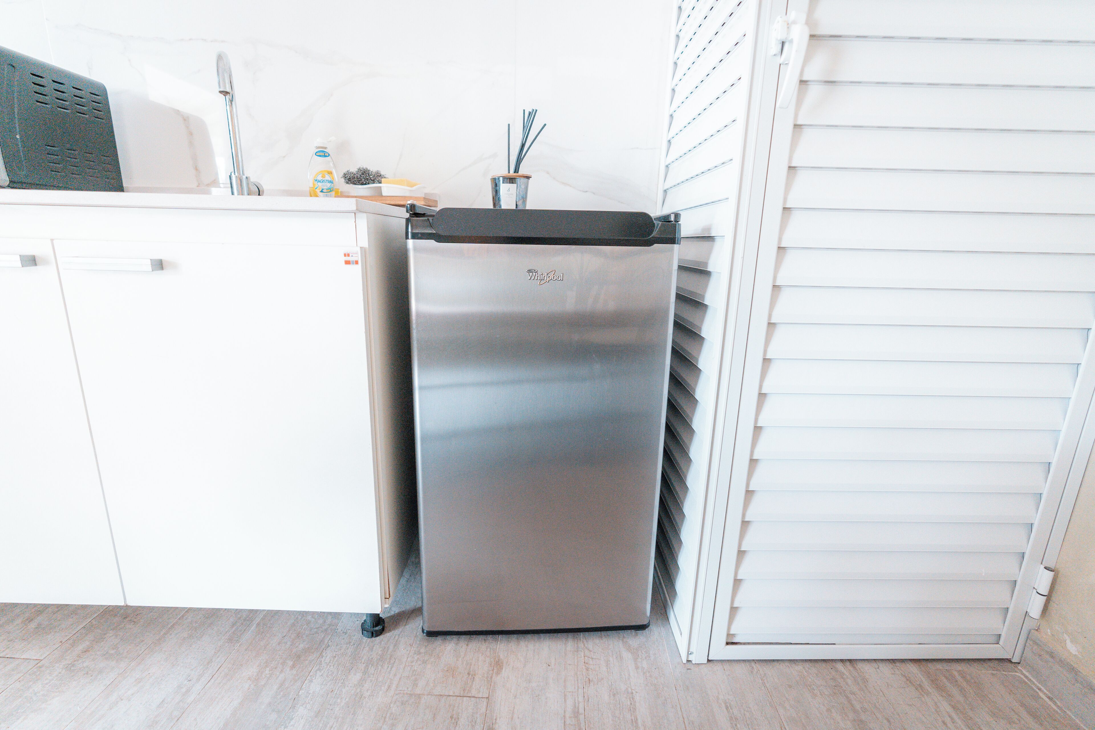 A compact kitchen with a stainless steel refrigerator, a white countertop, and a window with blinds.