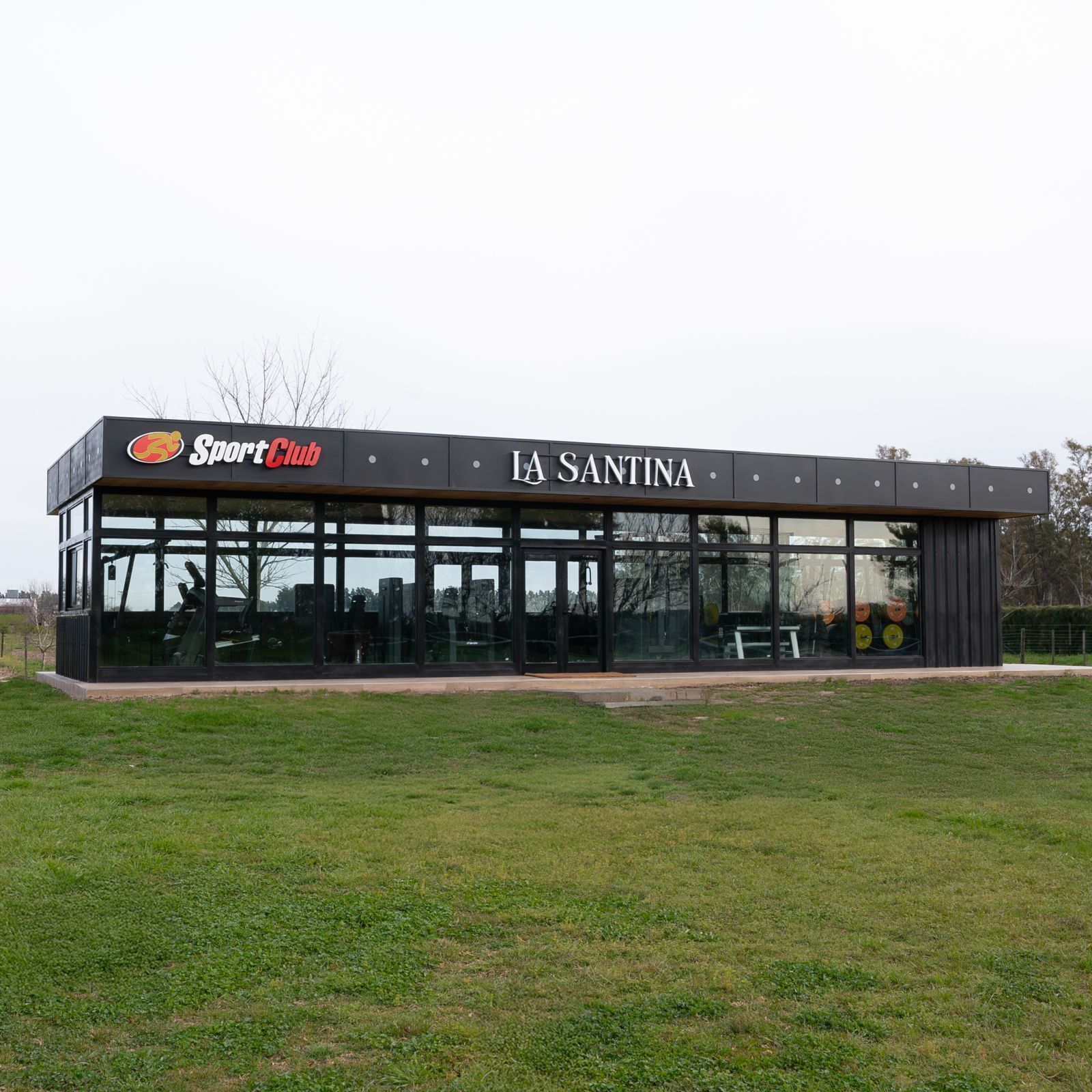 Un bâtiment moderne abritant un club sportif, doté de grandes fenêtres en verre et d’une enseigne indiquant : « Sport Club LA SANTINA ».