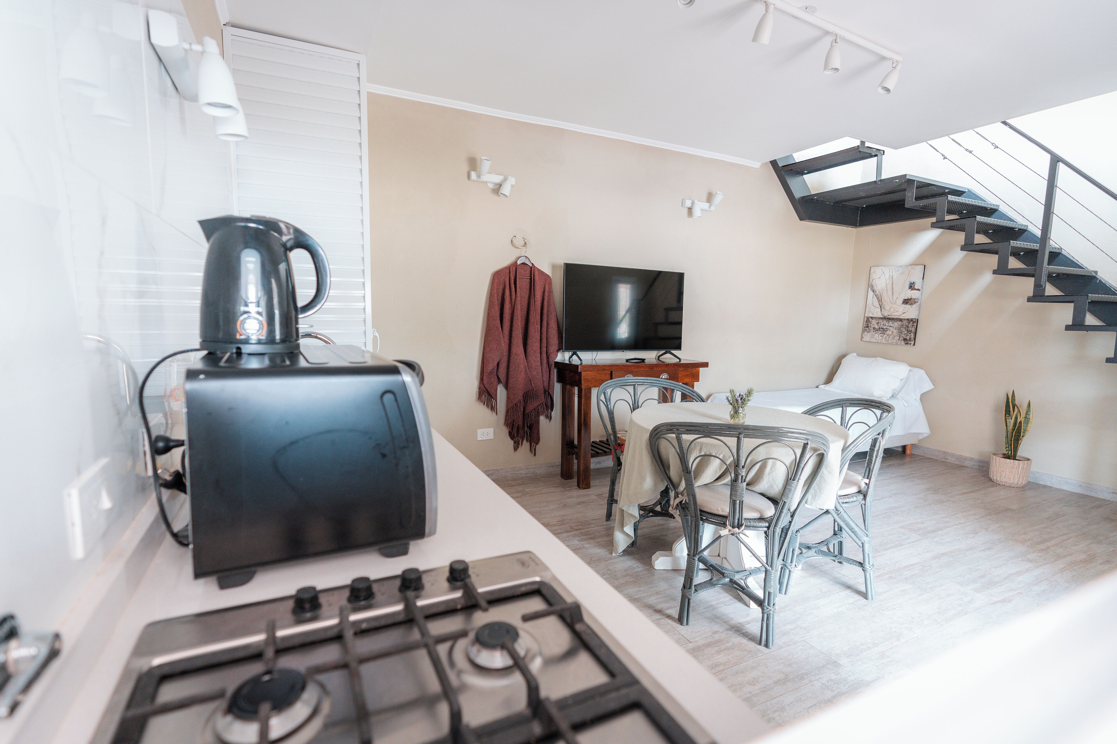 Una cocina moderna con estufa a gas, hervidor y tostadora. Hay un comedor con mesa y sillas, un televisor y una escalera.