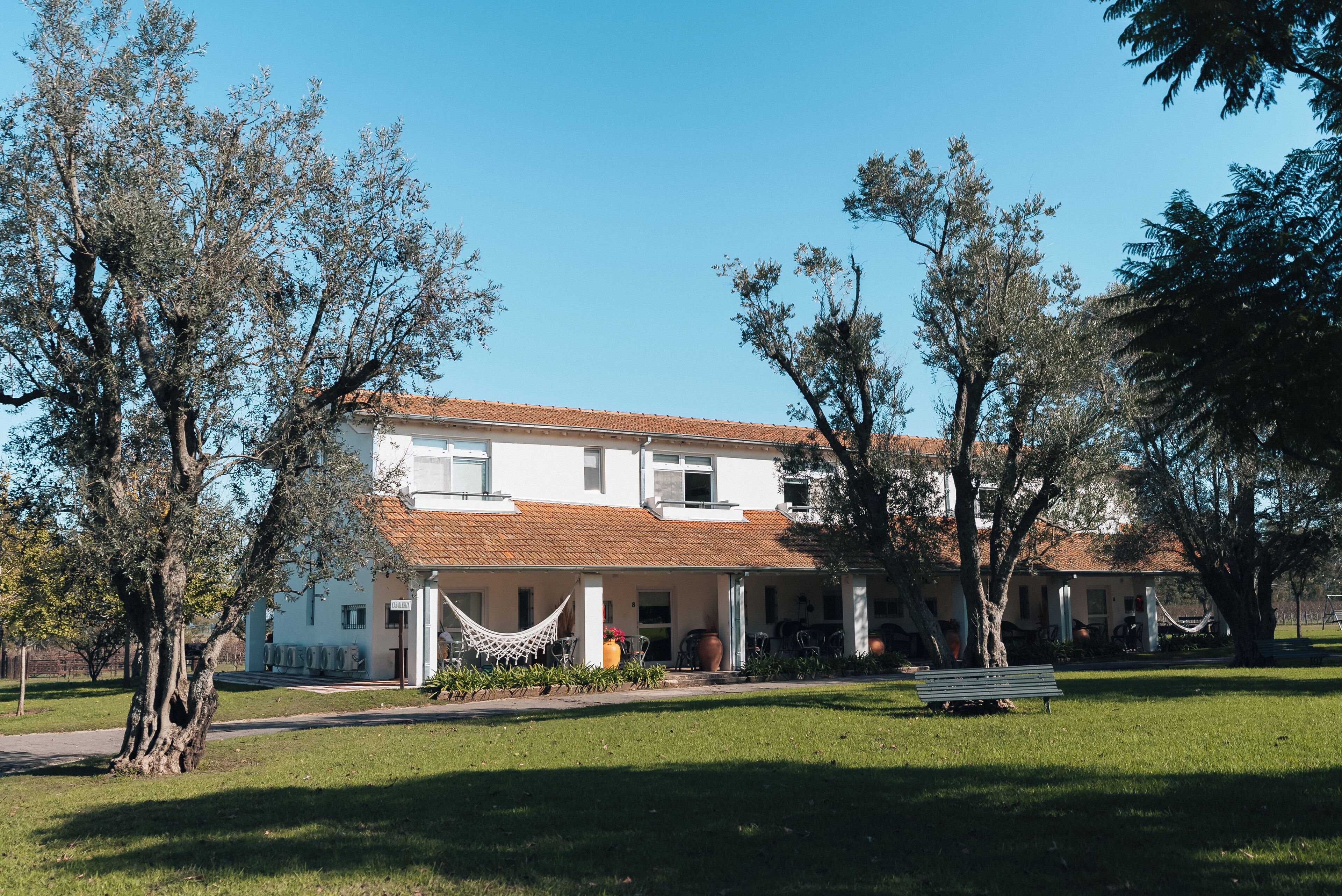 Un edificio blanco con techo de tejas, rodeado de árboles y un área verde con un banco.