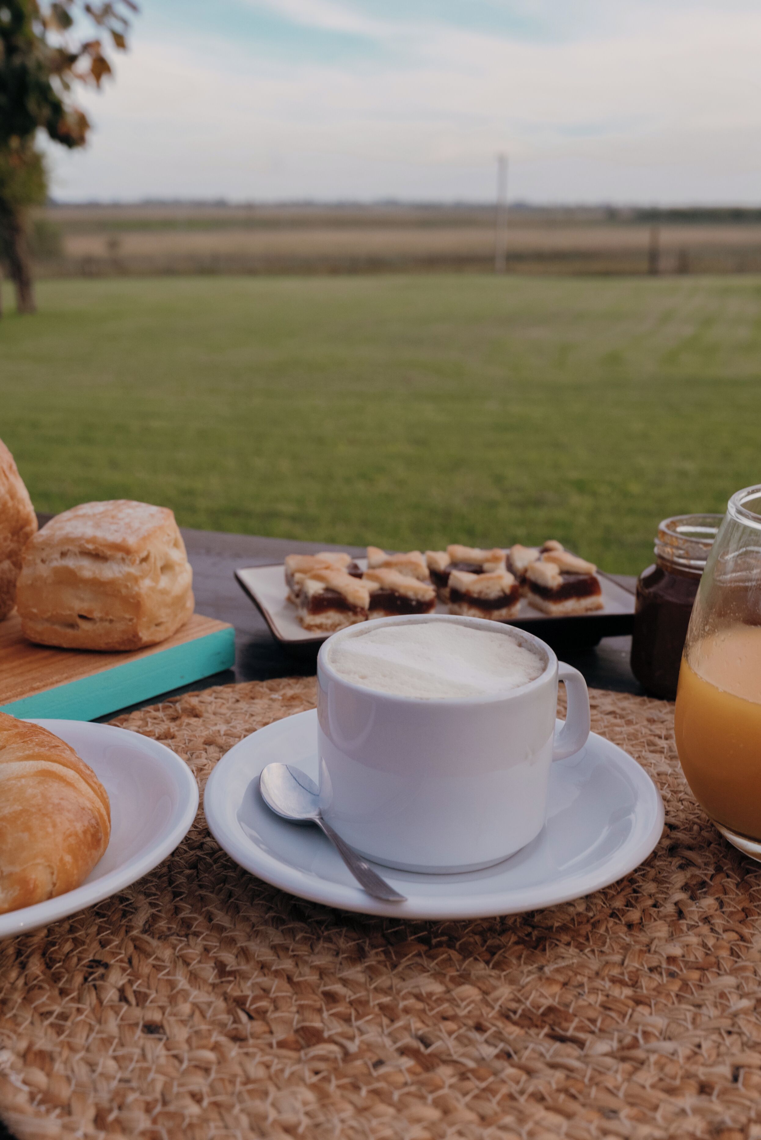A table with pastries, a cup of coffee, and a glass of orange juice, set outdoors with a grassy field in the background.