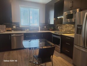 A modern kitchen with dark cabinetry, stainless steel appliances, a glass dining table, and a window with blinds. - Luxury Condo For Rent
 (Calgary)
