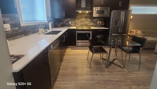 A modern kitchen with dark cabinets, a stainless steel refrigerator, a microwave, and a dishwasher. There is a window providing natural light and a small dining area with a table and chairs.