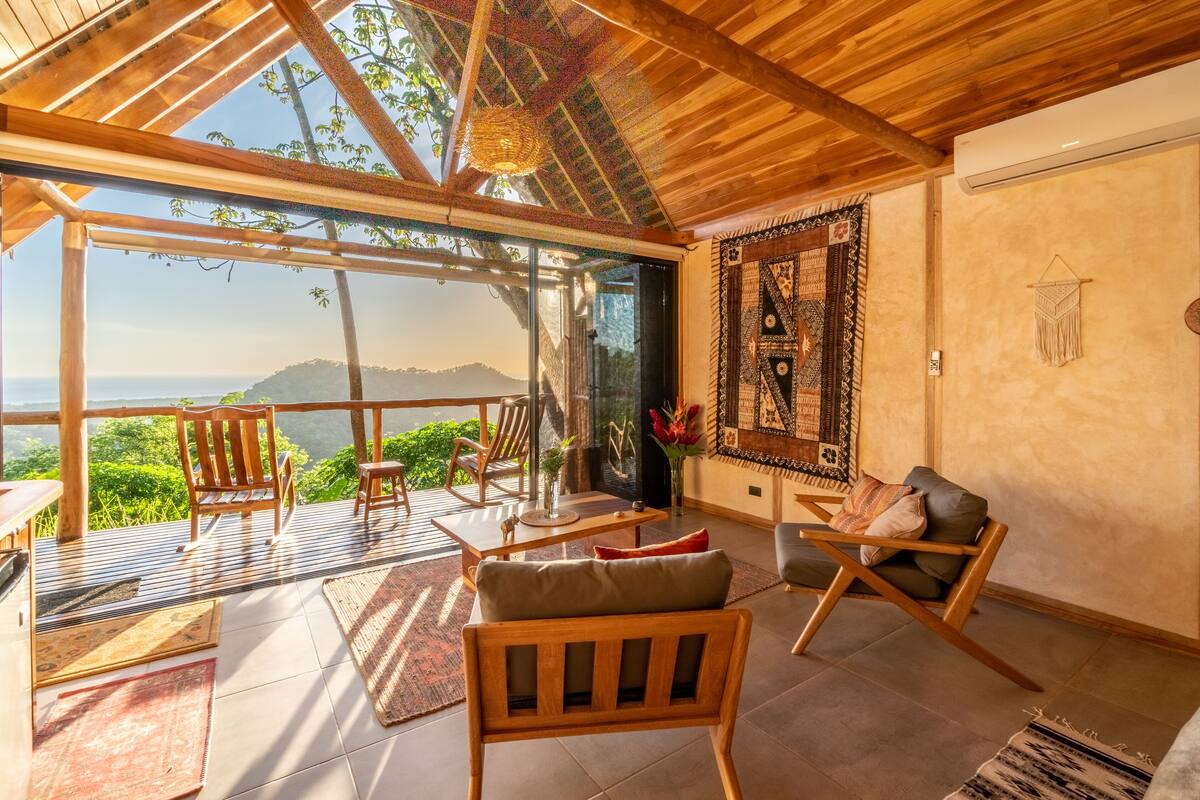 A spacious room with a large glass door leading to a balcony, wooden furniture, and a patterned wall hanging.