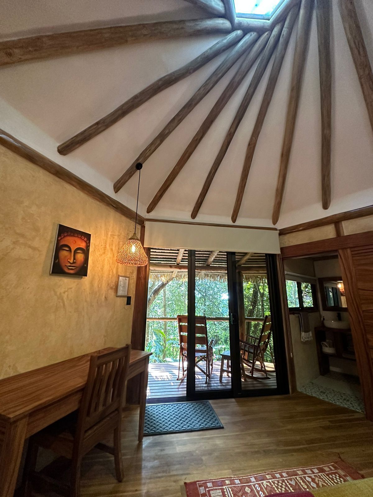 A room with wooden beams on the ceiling, a dining area with chairs and a table, and a view through a glass door to an outdoor area with chairs.