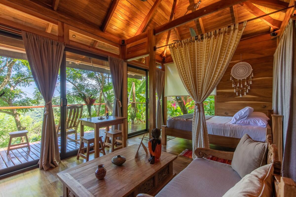 A wooden cabin with a bed, a table, and a chair. There is a view of greenery outside.