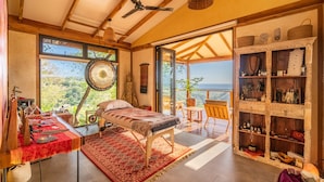 A room with a bed, a table with items, a shelf with decorative objects, and a view of the outdoors. - Tierra Encantada Wellness Center (Nosara)