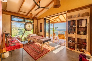 A room with a bed, a table with items, a shelf with decorative objects, and a view of the outdoors.