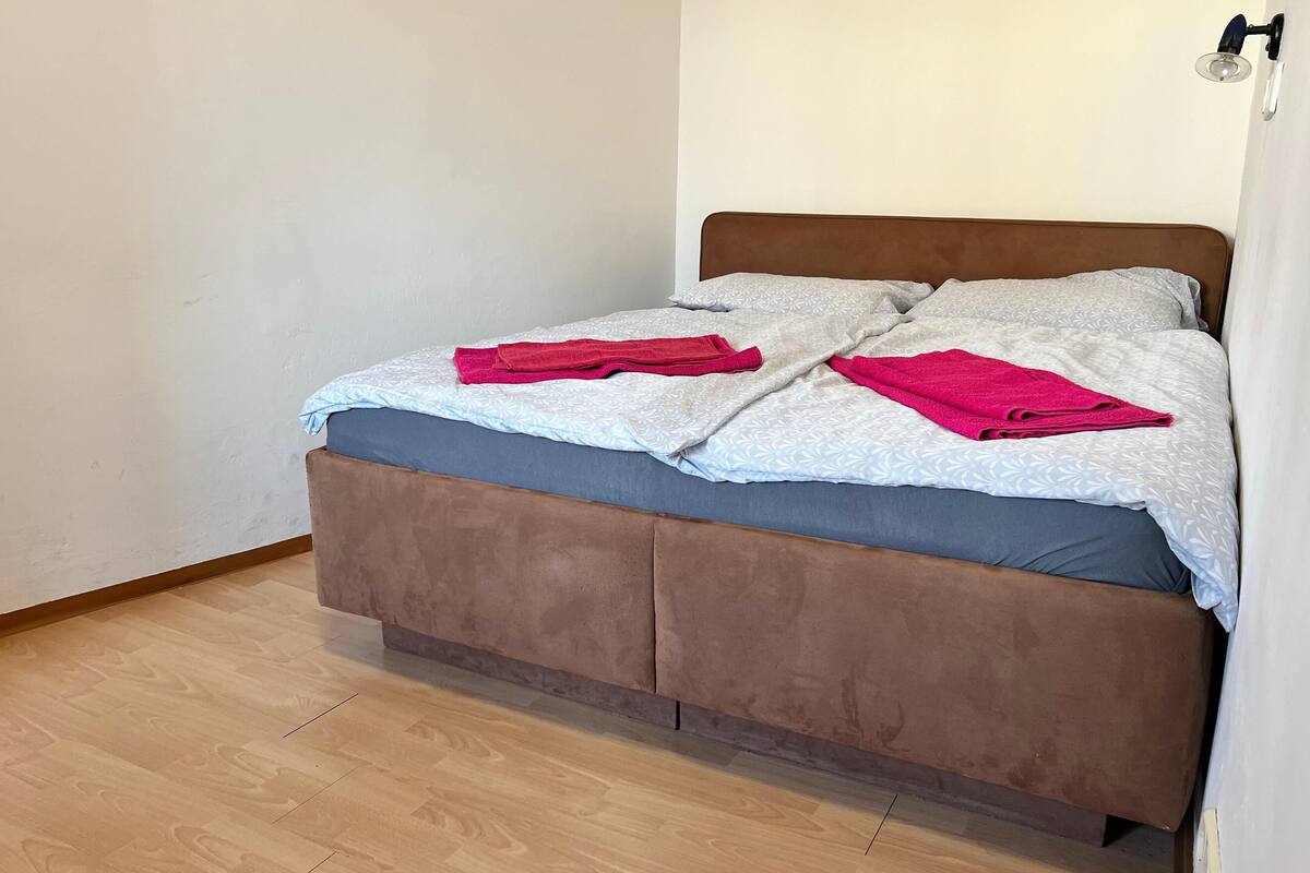 Uma cama com cabeceira marrom, roupa de cama estampada em cinza e branco, e duas toalhas vermelhas.