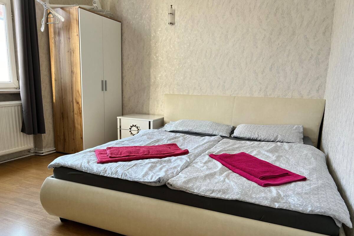 Quarto com cama, duas almofadas, mesa de cabeceira, guarda-roupa e janela com cortinas.