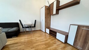 A modern living room with a wooden floor, a black sofa, a white table with two chairs, and built-in wooden cabinets. - EasyStay 3BR shared Apt. Near Meidling (Vienna)