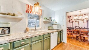 Una cocina con azulejos blancos tipo metro, un ventanal con cortinas, un lavabo, un microondas y una zona de comedor con sillas y mesa de madera.