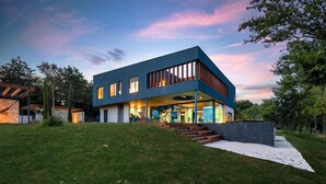 Una casa moderna de dos pisos con techo plano, ventanas amplias y un espacio exterior cubierto con asientos.