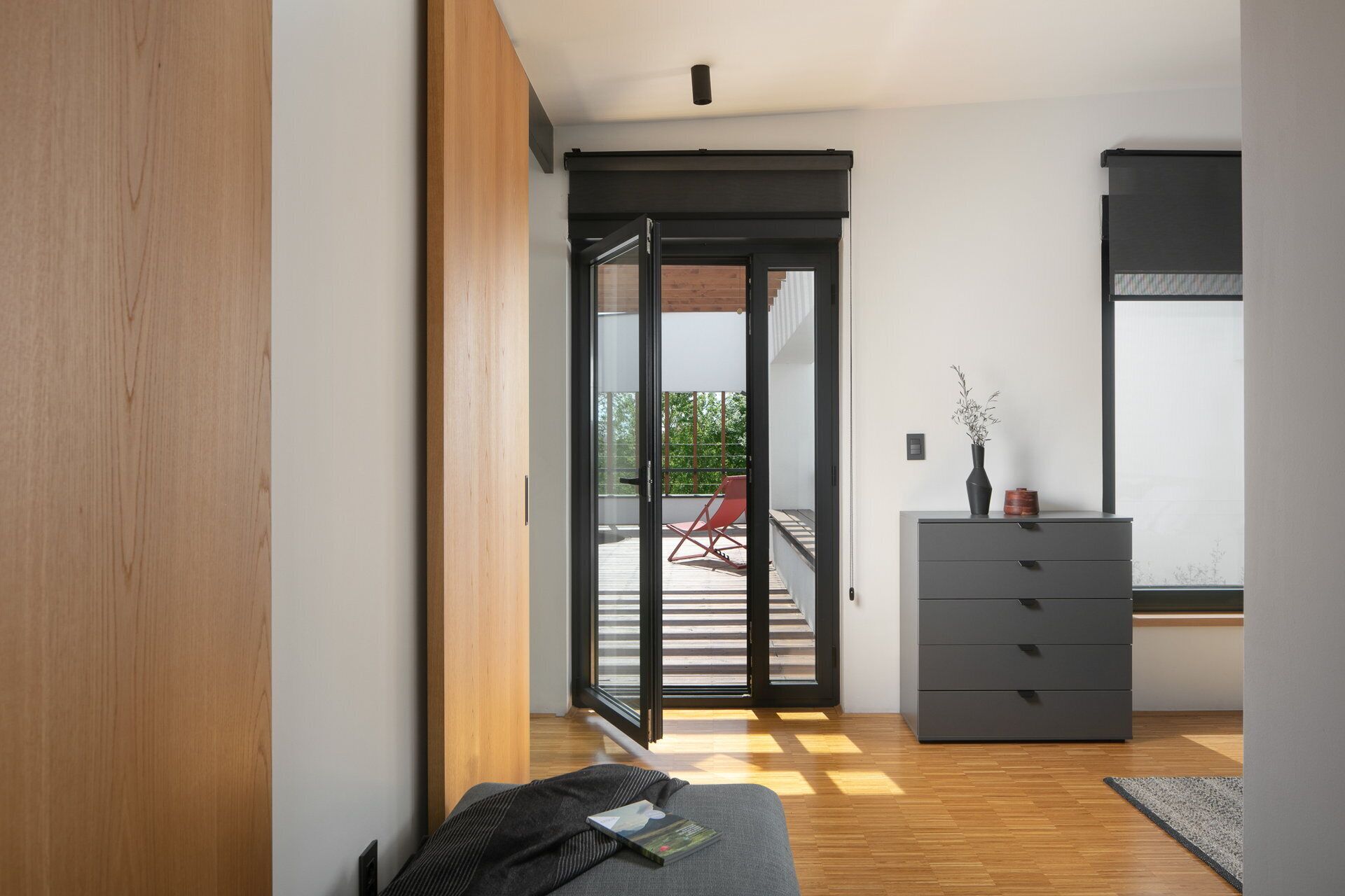 A modern interior with a wooden floor, a sliding glass door leading to an outdoor deck, a grey sofa, and a dark grey dresser with a vase.