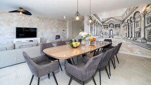 A modern dining area with a large table, grey upholstered chairs, and pendant lighting. A mural of classical architecture adorns the wall behind the table. - Beachfront Villa Atlantis with Pool (Kaštel Štafilić)