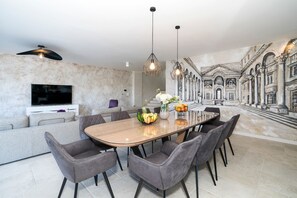 Ein moderner Essbereich mit einem großen Tisch, grauen Polsterstühlen und Pendelleuchten. An der Wand hinter dem Tisch ist ein Wandgemälde mit klassischer Architektur zu sehen.
