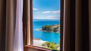 Blick auf die Küste durch ein Fenster mit Vorhängen, mit blauem Meer, felsiger Küstenlinie und Häusern mit roten Dächern.