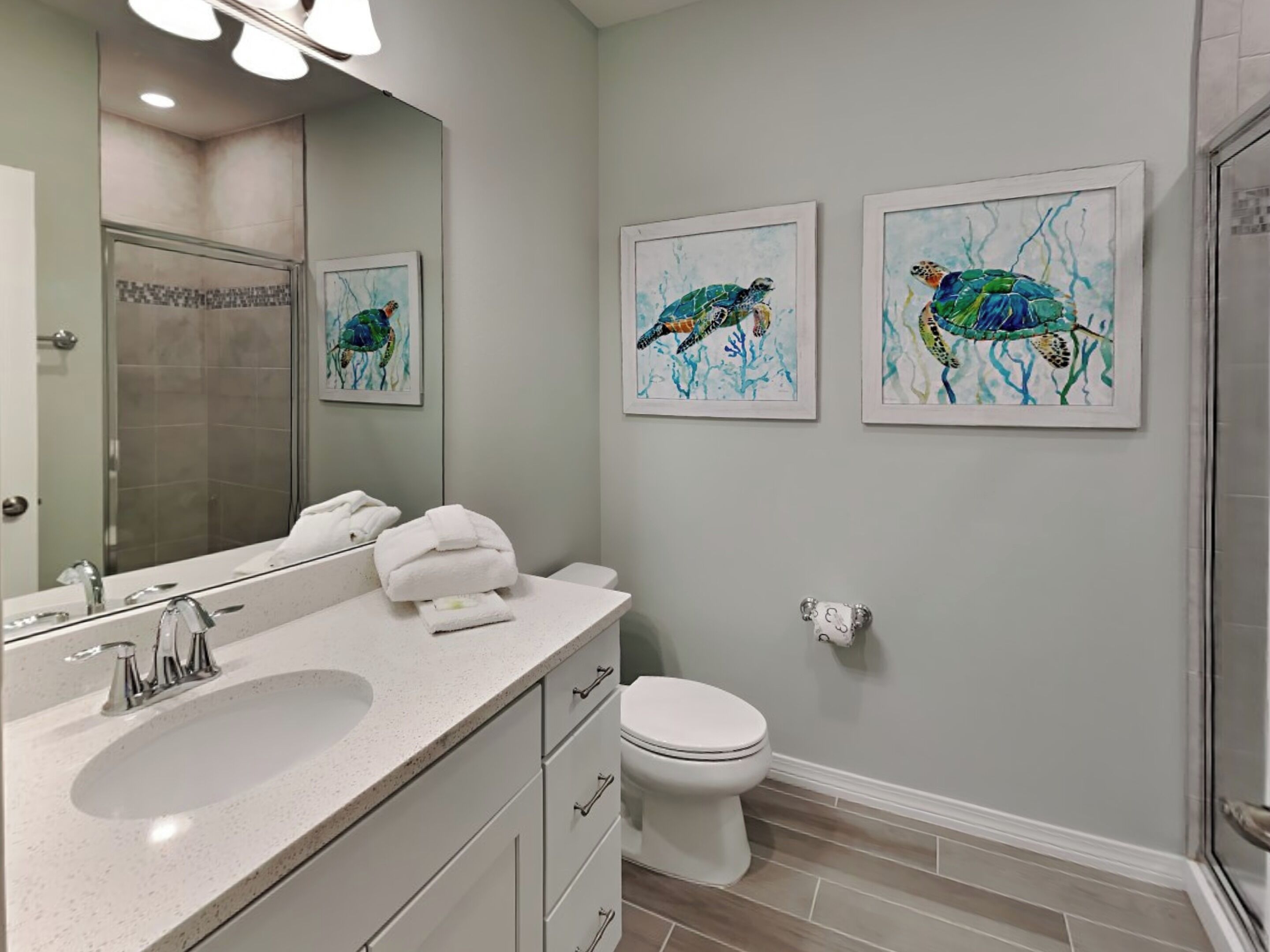 Une salle de bain équipée d’un lavabo, de toilettes et d’une douche. Deux tableaux encadrés représentant des tortues de mer sont accrochés au mur.