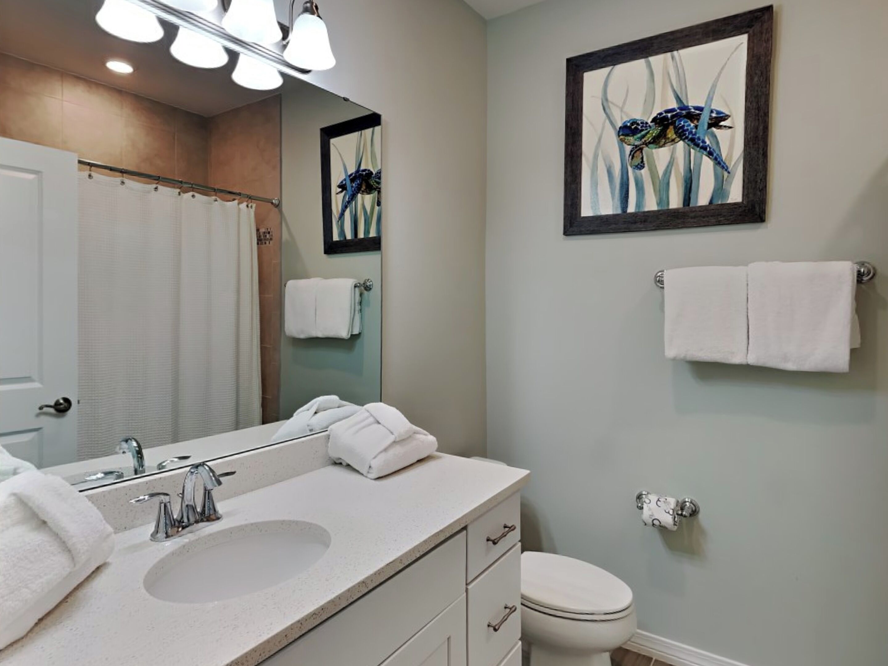 Une salle de bain avec un lavabo, un miroir et des toilettes. On y trouve un porte-serviettes avec des serviettes et une œuvre d’art encadrée accrochée au mur.