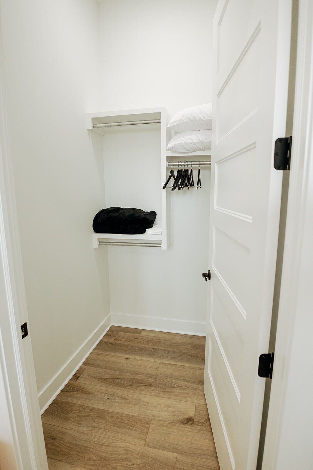 A small, narrow hallway with a wooden floor, a white door, and a closet with a hanging rack and shelves.