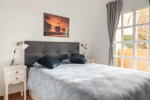 A neatly made bed with a grey headboard, two bedside tables with lamps, and a sunset painting on the wall.