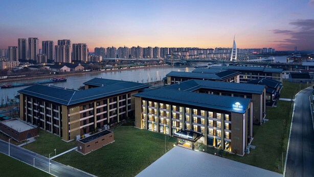 A modern hotel with multiple buildings, a river in the background, and a bridge. - Days Inn by Wyndham Yangzhou Guangling (Yangzhou)