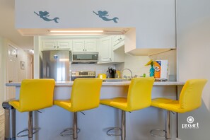A kitchen with white cabinets, a stainless steel refrigerator, a microwave, and a sink. There are four yellow bar stools with metal legs. - Hello Sunshine, 60 yards from the beach (Destin)