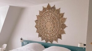 A bedroom with a bed, a teal nightstand, a wall hanging, and a white chair. - Hotel Liberté (Schoorl)