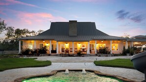 Una casa con patio de piedra, piscina con dos fuentes y un porche bien iluminado.
