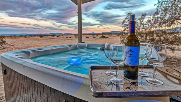 Un bain à remous avec une bouteille de vin et des verres sur une table, le tout installé dans un paysage désertique, avec un ciel au coucher du soleil en toile de fond.
