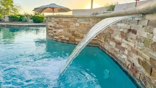 Una piscina con cascada, rodeada de muros de piedra y una sombrilla.