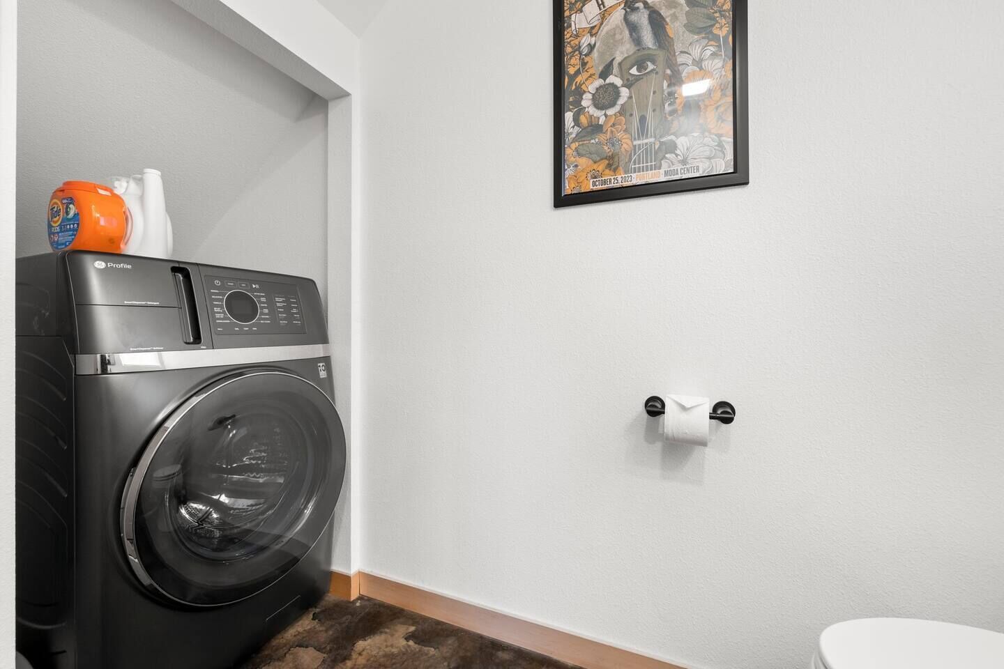 Un espace buanderie comprenant un lave-linge à chargement frontal, des toilettes et une œuvre d’art encadrée accrochée au mur.