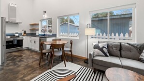 Eine moderne Küche mit weißen Schränken, ein Essbereich mit rundem Tisch und Stühlen, ein graues Sofa und ein großes Fenster mit Blick auf ein blaues Haus und einen weißen Zaun.