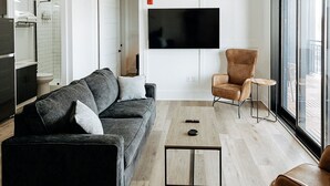 Una sala de estar moderna con un sofá negro, un sillón marrón, una mesa de centro y un televisor montado en la pared.