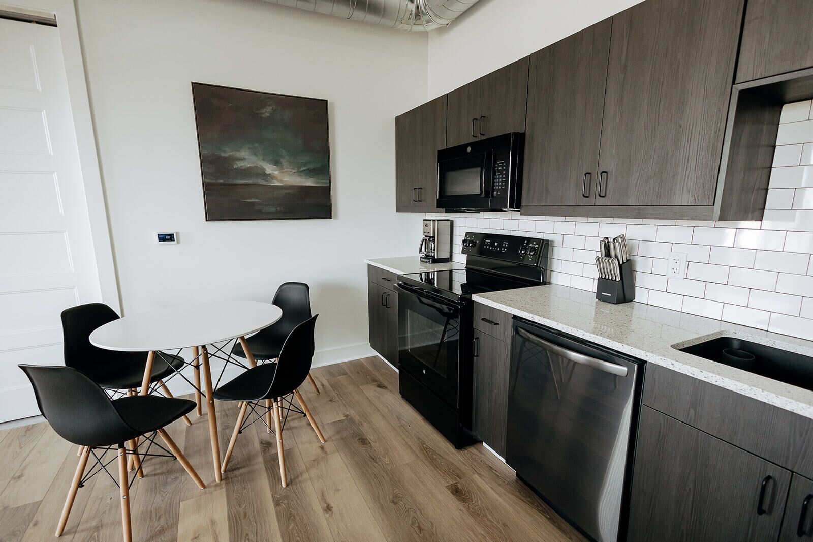 A modern kitchen with dark wood cabinets, a stainless steel microwave, stove, and dishwasher. There is a white countertop, a small dining table with black chairs, and a painting on the wall.