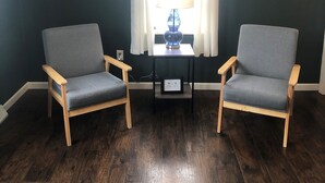 Une pièce avec deux fauteuils gris, une petite table avec une lampe et une fenêtre ornée de rideaux blancs.