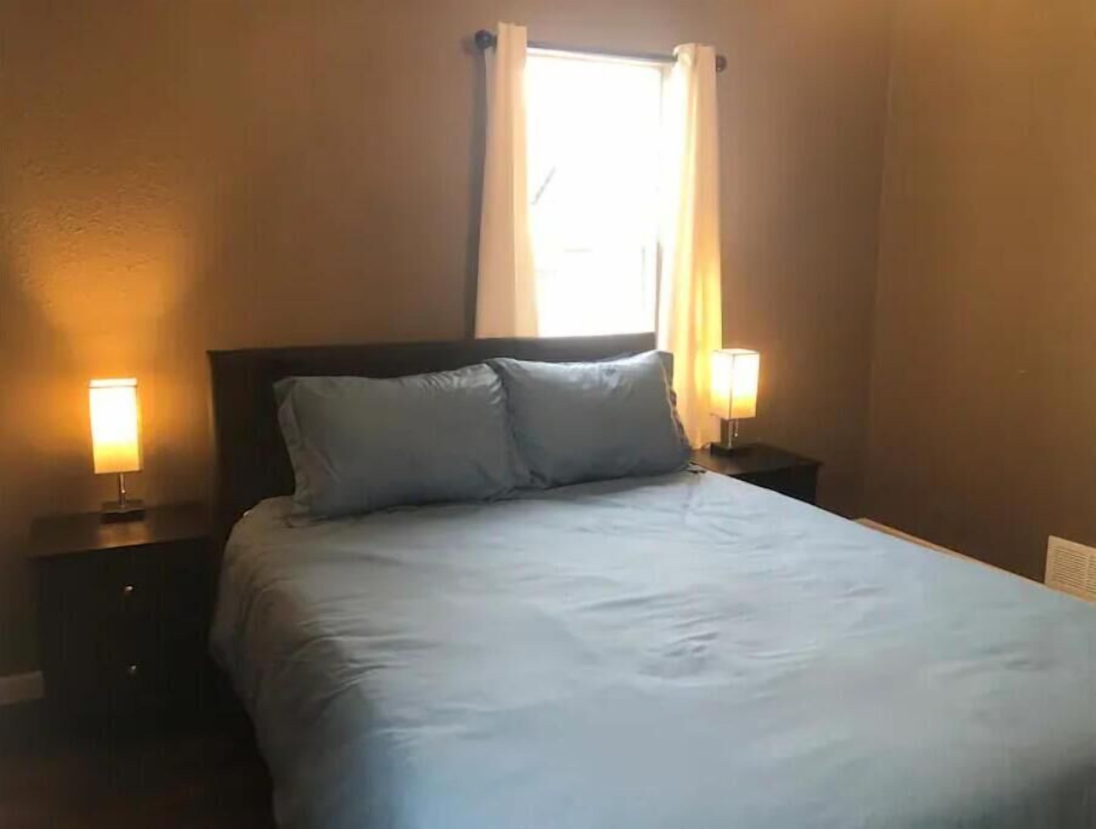 Una cama bien hecha con dos almohadas, una ventana con cortinas y dos lámparas de noche a cada lado.