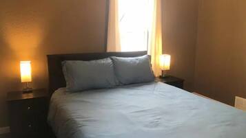 Una cama bien hecha con dos almohadas, una ventana con cortinas y dos lámparas de noche a cada lado.