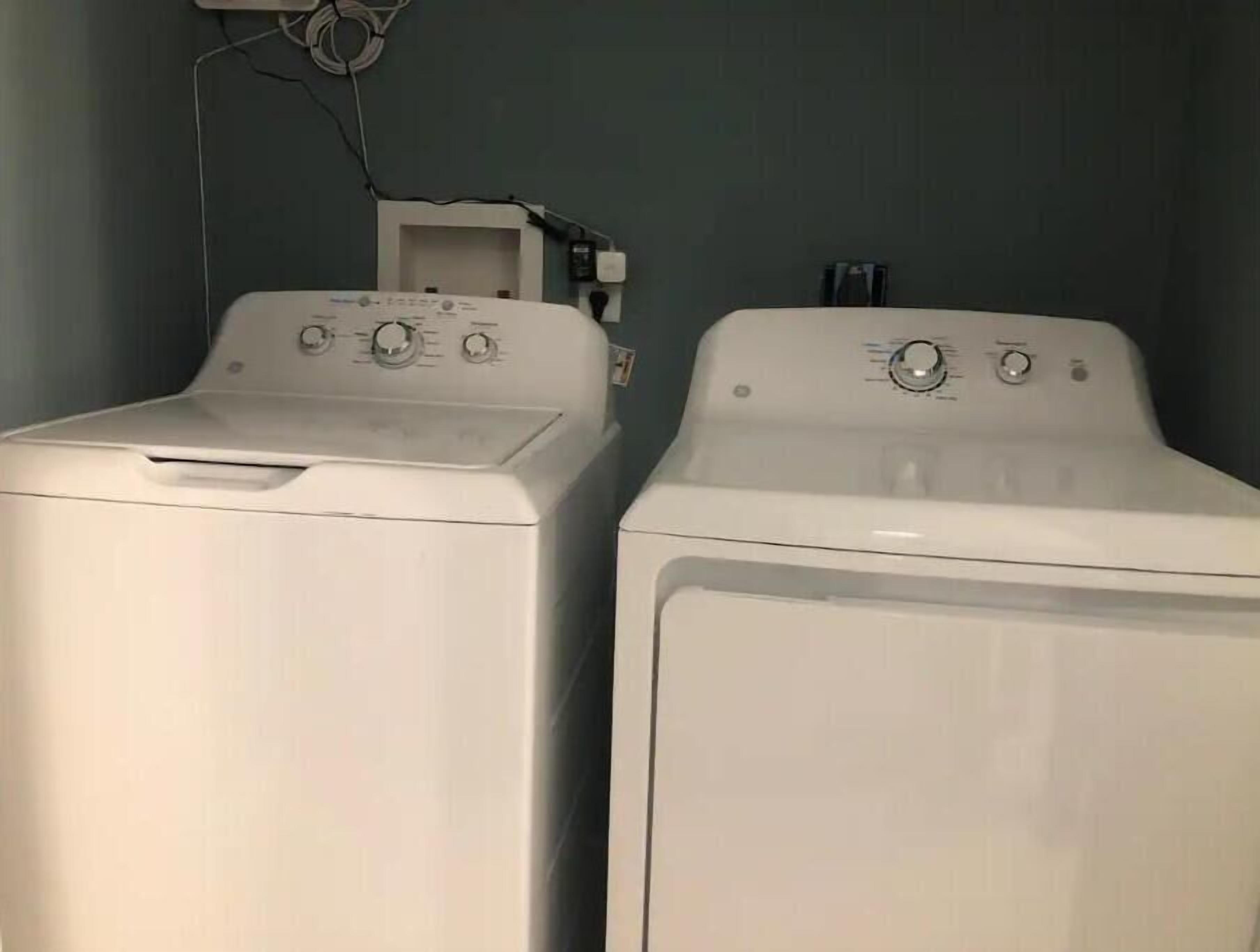 Two white front-loading washing machines in a laundry room with a plain wall and a power outlet visible.