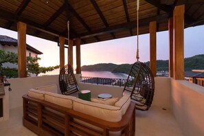 A covered outdoor seating area with a wooden bench and two hanging chairs, overlooking a body of water and hills.