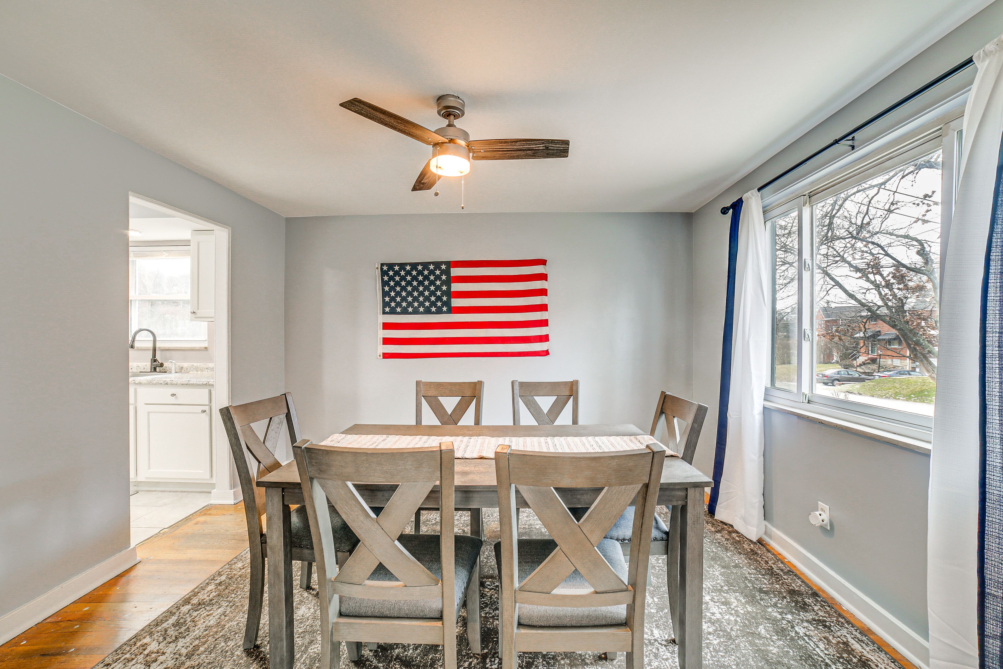Uma sala de jantar com mesa e cadeiras, um ventilador de teto, uma janela com cortinas e uma bandeira americana na parede.
