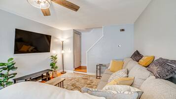 Een moderne woonkamer met een bank, een aan de muur bevestigde televisie, een plafondventilator en een trap naar een ander niveau.